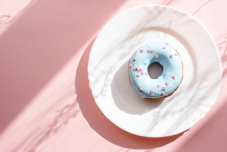 甜甜圈覆盖着蓝色的釉面, 洒上粉红色背景上的白色盘子上的小粉红色的心与表面图案的阴影