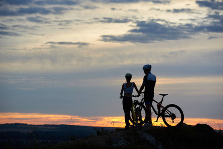 两个骑山地自行车的年轻人站在悬崖顶上, 日落时有美丽的山丘和天空景色。健康生活方式的概念