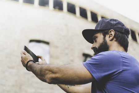 胡子肌肉男子身穿黑色 snapback 帽, 并使自拍与他自己的智能手机。在街上行走和使用手机的人。背景模糊。水平