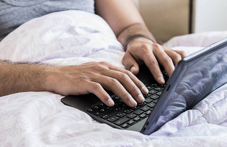 在床上在键盘上打字的笔记本电脑上工作的年轻男性手手指的特写