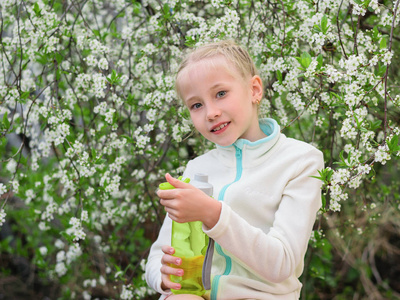 穿着运动服的女孩在樱花里捧着一瓶清爽的饮料。樱背景下的女孩肖像