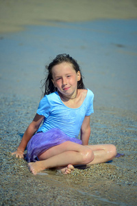 一个穿着紫色泳装的可爱小女孩坐在沙滩上的湿沙子上, 阳光明媚的一天, 看着相机, 微笑着。图像的垂直方向