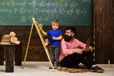 老师看着孩子擦黑板。坐在地板上的微笑的人和严肃的面孔站立在他旁边的孩子