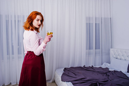 华丽的红头发的女孩在粉红色的上衣和红色的裙子与酒杯在手边的房间附近的床上