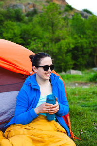 在森林里露营。这个女孩正在从帐篷附近的一个热杯子里喝咖啡, 在山的背景下。一个女人坐在帐篷旁边的睡袋里喝咖啡