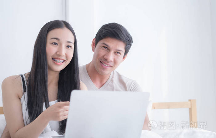 快乐的亚洲情侣用膝上型电脑在晨床上微笑