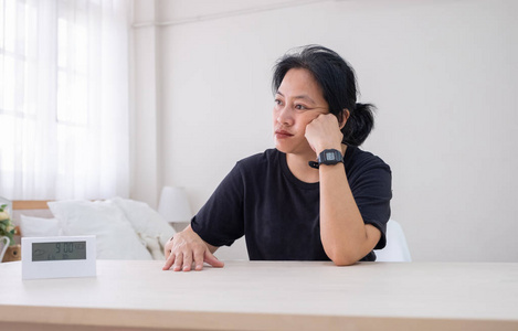 坐在木桌旁的亚洲女子在白色卧室里用数字钟在手上放着忧郁的情绪. 等待时间不太好
