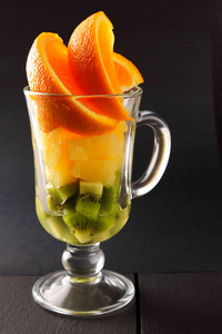在玻璃杯中的热带水果片。水果鸡尾酒在黑色的背景。橘子片, 猕猴桃, 玻璃碗里的菠萝片。柑橘类水果