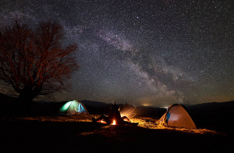 在山上露营的夜晚。两个年轻的游客, 男孩和女孩坐在两个帐篷之间的剪影在燃烧篝火背景下的黑暗星空。旅游和旅游概念