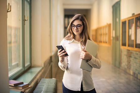 快乐的年轻女人在大学的走廊里检查她的智能手机