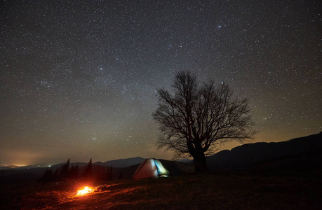 明亮的篝火在旅游照明帐篷附近燃烧。夜露营在山下美丽的星空。背景上的大树和远处山丘的剪影。旅游业和旅游概念