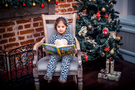 漂亮的小女孩微笑着靠近圣诞树坐在老式椅子上的玩具熊。新年快乐