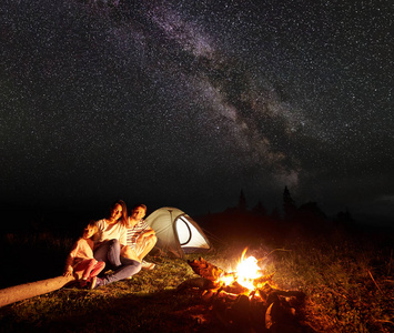 家庭露营在山上的夜晚, 坐在一个日志前的照明帐篷和燃烧篝火下美丽的星空和银河, 微笑着相机。旅游业和旅游概念