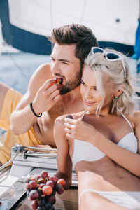 微笑的年轻夫妇在泳装吃葡萄在游艇上