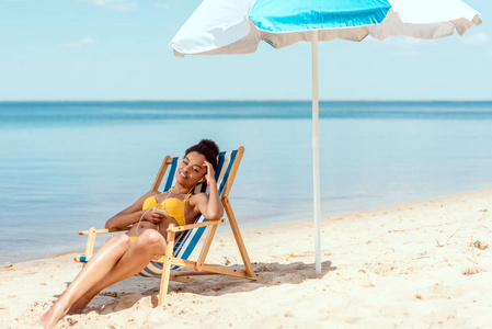 年轻的非洲裔美国妇女放松在甲板上的椅子和听音乐耳机与智能手机下海滩伞在海前
