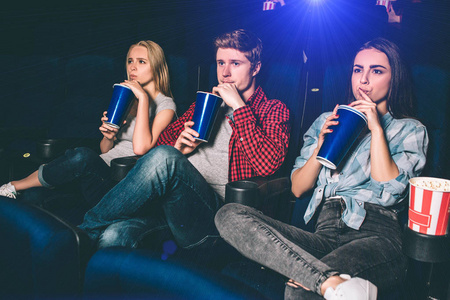 三朋友坐在一起, 同时喝可乐。他们正在看电影。年轻人是认真的, 专注于观看