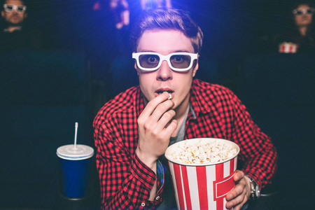 他坐在电影院大厅的椅子上吃爆米花的画像。他看上去很紧张。他戴着特别的眼镜看3d 部电影。他还吃着篮子里的爆米花