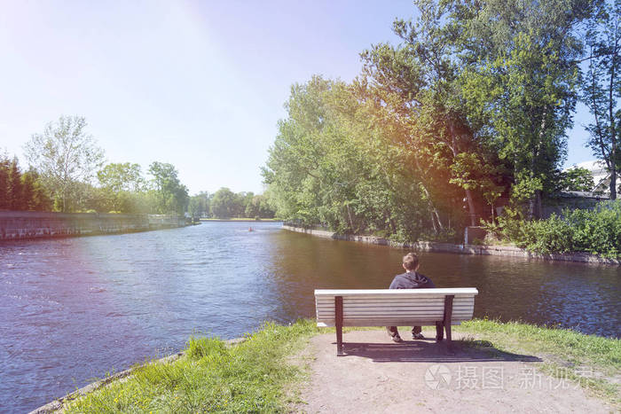 一个年轻人坐在河边的长凳上看着那艘运动船照片-正版商用图片0ost07