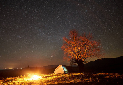 梦幻般的夜露营网站视图。明亮的篝火在美丽的星空下在旅游照明帐篷附近燃烧。大树和远处山脉的背景。旅游业和旅游概念