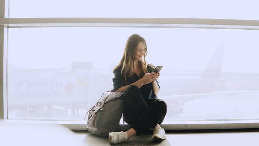 快乐的女人坐在智能手机的机场窗口。高加索女孩与背包使用信使应用程序在终端。4k
