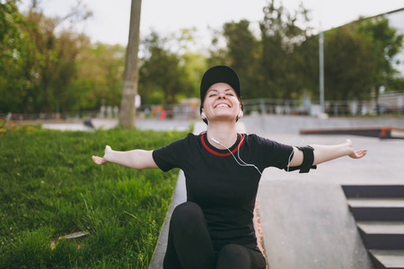 年轻运动放松的女孩在黑色制服与耳机听音乐, 传播手, 休息和坐在运行之前或之后, 在城市公园户外训练。健身, 健康的生活方式