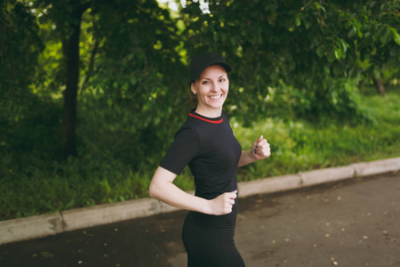 年轻的微笑运动美丽的黑发女孩在黑色制服和帽训练做运动演习, 运行和看在城市公园的道路上的户外的相机。健身, 健康的生活方式概念