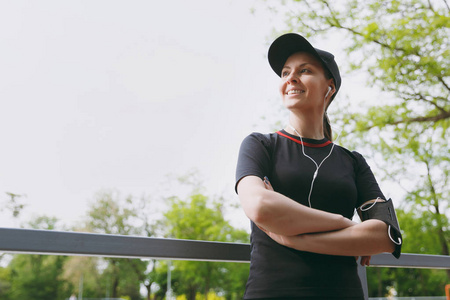 年轻的运动微笑的女孩身着黑色制服, 帽子与耳机听音乐, 站立保持双手折叠前或后运行, 在城市公园户外训练。健身, 健康的生活方式