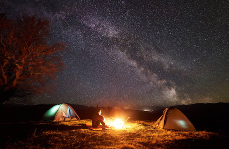 晚上露营在山上。徒步旅行者坐在草地山谷两个照明帐篷之间, 享受着明亮的燃烧篝火。大树和令人惊叹的星空, 银河的背景