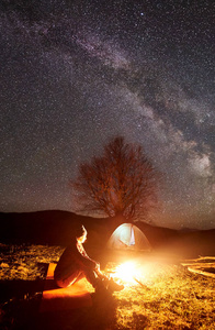 在山上露营的夜晚。明亮地点燃了燃烧的篝火黑暗的剪影坐在黑暗的星空下的年轻女孩与旅游帐篷和遥远的丘陵背景。旅游和旅游概念