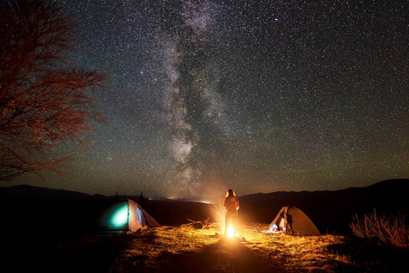 晚上露营在山上。女性徒步旅行者的剪影站立在两个发光的旅游帐篷之间在燃烧的篝火附近大树, 享受惊人的星空和银河。户外活动概念
