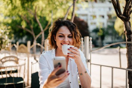 一个微笑的年轻妇女的肖像喝咖啡和使用智能手机户外