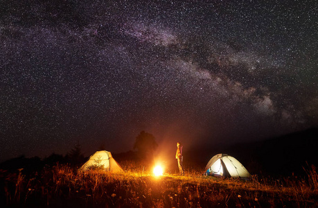 在山上露营。年轻苗条的旅游女孩站在两个帐篷之间看着明亮的燃烧篝火在深黑的天空与许多明亮的闪闪发光的明星背景。旅游和旅游概念