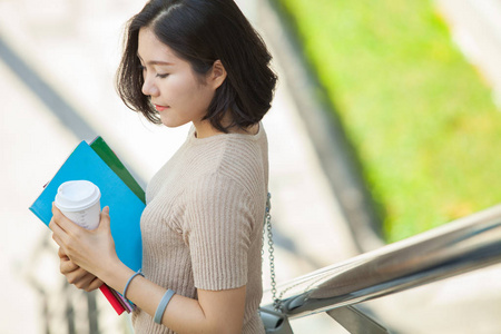 美丽的亚洲女大学生拿着她的书和一杯咖啡站在户外, 人们教育学习高中计划聪明的青少年概念