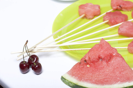 西瓜的果肉是用邮票切碎的。它们被赋予不同的形式。串在一起野餐。旁边的盘子是一块西瓜和一些甜樱桃浆果