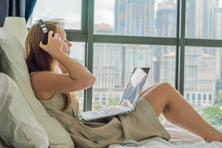 年轻女子正在他的床上的笔记本电脑上的背景下, 俯瞰摩天大楼的全景窗口。使用无线耳机。自由职业者, 远程工作, 在家工作