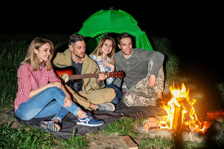 年轻人在晚上篝火旁为朋友弹吉他。野营季节
