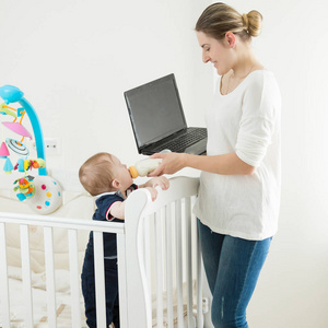在笔记本电脑上工作的年轻的自我 emploed 妇女的肖像, 同时喂养她的宝宝从瓶子牛奶