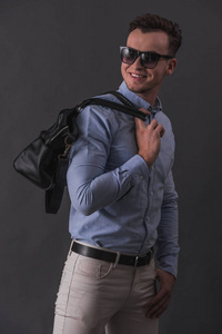 时尚的年轻商人在智能休闲服装和眼镜拿着一个袋子和微笑, 在灰色背景