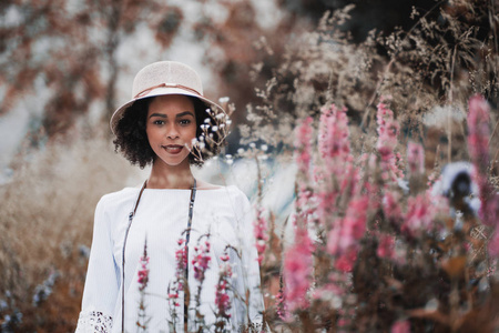 一个年轻快乐的黑色旅游女性的肖像在帽子和衬衫站在夏天的田野上, 周围五颜六色的乡土花草和花, 看着相机场浅景深