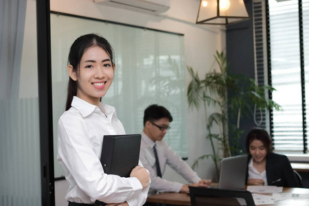 领导企业女性理念。开朗年轻的亚洲女商人与环活页夹站在她的同事在办公室背景