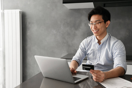 一个微笑的年轻亚洲男子的肖像手持塑料信用卡, 而坐在桌子上的笔记本电脑在家里