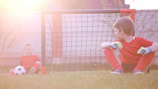 小足球运动员正坐在草坪上的足球球门上。