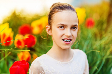 春天的可爱的小女孩，穿着礼服，坐在户外用黄色和红色的郁金香，在背景上的画像。青春期女孩戴着牙套的牙齿