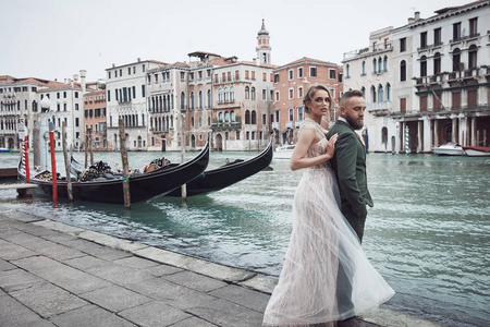 在威尼斯运河附近摆着一对新婚夫妇。优雅的女人在奢华的象牙礼服, 凌乱的发髻头发, 男人在绿色的三件西装
