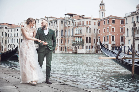 迷人的新婚夫妇在威尼斯运河附近散步。优雅的女人在奢华的象牙礼服, 凌乱的发髻头发, 男人在绿色的三件西装