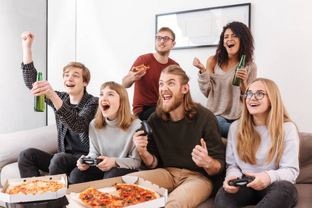 一群快乐的朋友坐在沙发上, 尖叫着, 在一起消磨时间, 玩电子游戏。年轻人在家里吃比萨饼和喝啤酒的画像