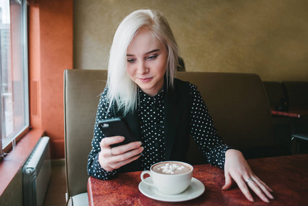 年轻漂亮的金发女孩看着电话，坐在咖啡馆里。咖啡和果汁软糖吃早饭