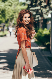 年轻的微笑的女人在时髦的服装与智能手机在街上的侧面视图