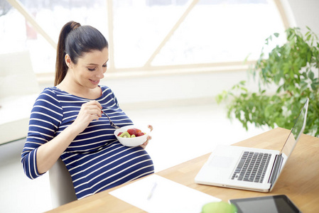 美丽的年轻孕妇吃一碗水果沙拉, 而坐在笔记本电脑前和在家放松