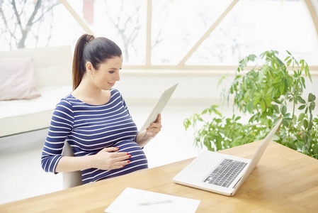 美丽的孕妇使用数码片和笔记本电脑, 而坐在办公桌在家里